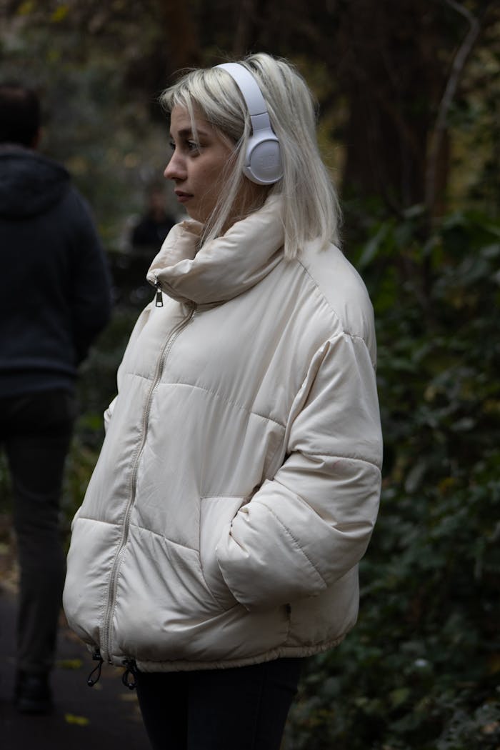 about-us A blonde woman with headphones enjoys music outdoors in a white puffer jacket, winter setting.